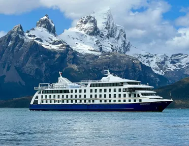 Wide shot of cruise ship, craggy mountains, clouds.