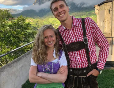 Two locals smiling, large mountains, clouds behind them.