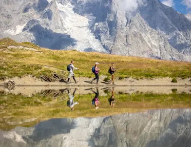Three guests walking past reflective lake, meadow, snowy mountain in distance. 