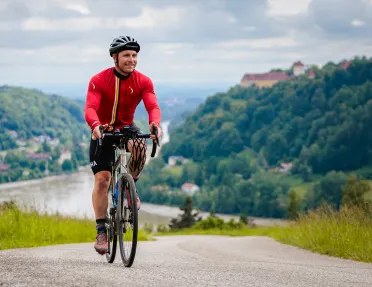 Man riding a bike on an asphalt road, with mountains of trees behind him