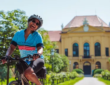 Woman wearing sunglasses and riding a bike in front of a long pathway leading to a yellow building