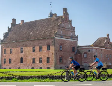 Two bikers in front of a large red building in the middle of a grass field