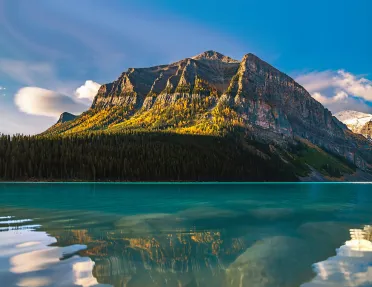 Lake in front of tall trees and a tall mountain, with the sunshine hitting the mountain