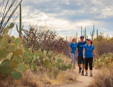 Two women and one man hiking on a dirt trail surrounded by cacti and dried trees