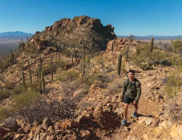 Man ascending a rocky, dirt trail with canyons and cacti behind him