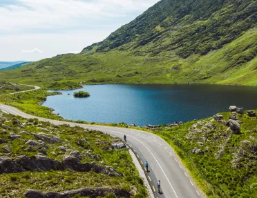 Road surrounded by large grass hills and a clear, blue lake to the right