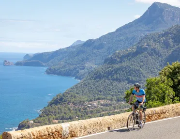 Man riding a bike on a road, looking out to the ocean and a cliff covered with trees