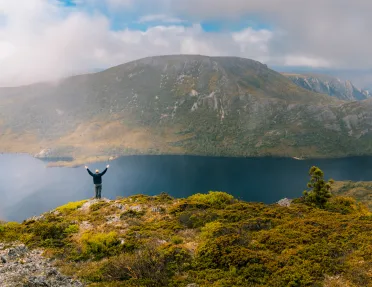 Man with his arms open while standing on a cliff, looking out to a large lake