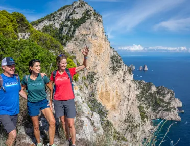 Two women and a man smiling and waving, while standing on a cliff next to the ocean