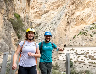 Man and woman walking on a wooden bridge with a large cliff in the background