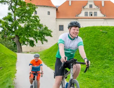 Two men smiling while riding bikes on a road, with a beige and orange castle-like building in the back