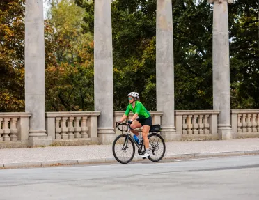 Woman wearing green biking gear, riding a bike on a road with large stone pillars in the background