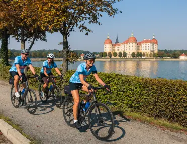 Three bikers on a road with a lake and large building in the background