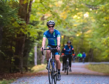Man wearing sunglasses, riding a bike on a road surrounded by tall trees