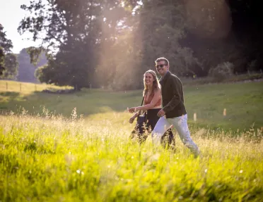 Man and woman smiling while walking through a grass valley, with trees and tall weeds