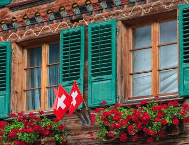 Wooden cabin building, with three Swiss flags and flower beds under a window