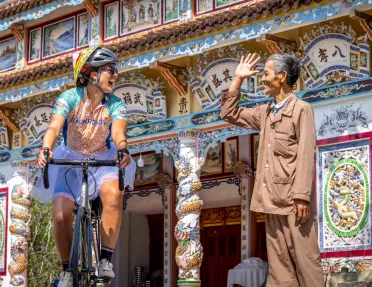 Woman riding a bike in front of a large temple, with a man smiling and waving at her