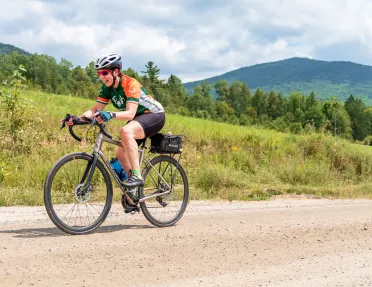 Woman riding a bike on a dirt road, with a grassy hill in the background