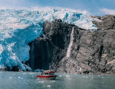 Red and gray boat floating next to large glaciers and cliffs