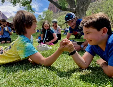 Two kids laying down on the grass, arm-wrestling