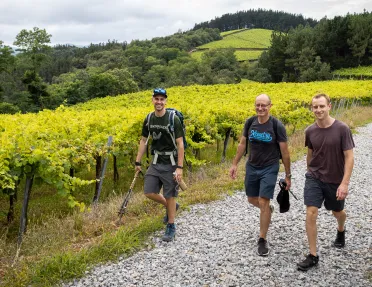 Three men smiling while hiking on a gravel trail, with crop fields in the background