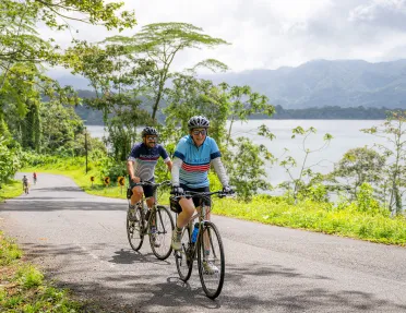 Two people riding bikes on a road, with a lake to the right
