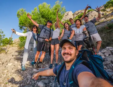 Group of men and women, holding hiking poles and smiling with their arms open on a rocky trail