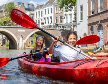 Two girls paddling in a red kayak on a river, with a town in the background