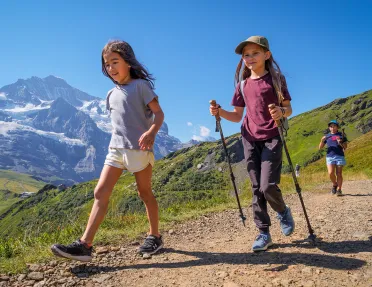 Two girls with hiking poles, walking on a gravel trail with large mountains in the distance