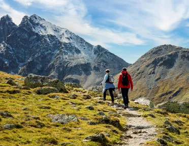 Man and woman ascending a rocky, dirt trail with large mountains ahead
