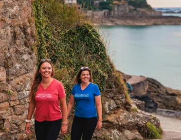Two women walking along a stone back with hills and the ocean in the background