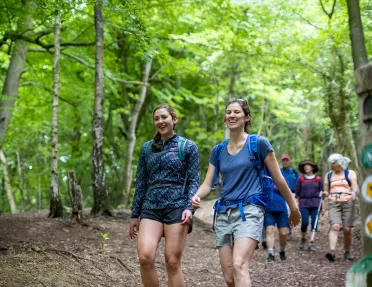 Two women walking along a dirt trail with tall trees in the background
