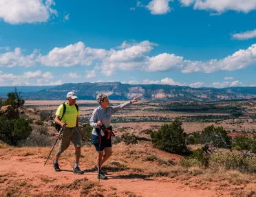 Man and woman with walking poles on a dirt trail, pointing out to tall mountains