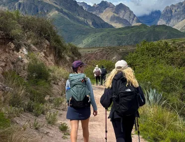 Two women walking on a dirt trail, with large hills and mountains in the distance