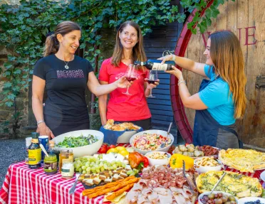 Three women behind a table full of food, pouring wine into glasses