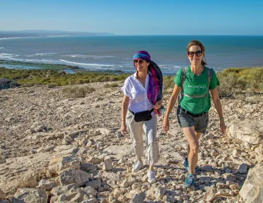 Two women smiling while walking on a gravel trail with the beach in the background