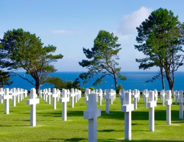 Rows of white cross-shaped headstones stretch across a manicured lawn at a coastal military cemetery, with pine trees and the blue sea visible in the background.