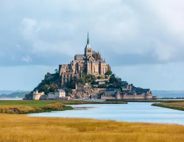 Mont Saint-Michel rises above surrounding tidal flats and waterways, showcasing its medieval abbey, stone ramparts, and historic village beneath a cloudy sky.