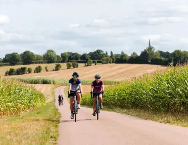 Two cyclists ride along a quiet country road bordered by tall cornfields, with rolling farmland, hedgerows, and a distant church spire stretching across the rural landscape.