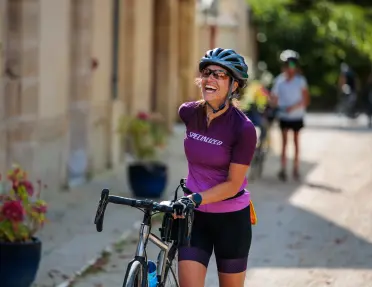 Cyclist standing beside a road bike on a village street lined with historic stone buildings and potted flowers, with other cyclists visible in the background.