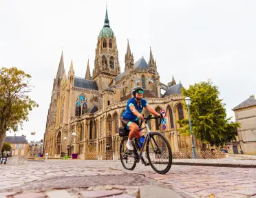 Cyclist rides across a cobblestone square in front of a Gothic cathedral featuring soaring spires, stained-glass windows, and ornate stone architecture in a historic French town.