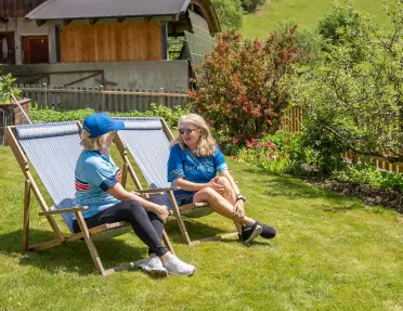 Two cyclists relax in reclining lawn chairs in a landscaped garden, surrounded by flowers, trees, and alpine countryside. Traditional wooden buildings and green hills form the backdrop.