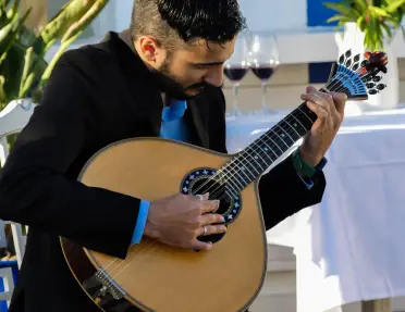 Musician playing a traditional Portuguese stringed instrument at an outdoor dining venue. White table linens, wine glasses, and potted plants create an elegant setting, while sunlight illuminates the performance area and the instrument's detailed craftsmanship.