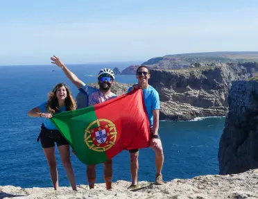 Three travelers stand on a dramatic coastal cliff overlooking the Atlantic Ocean, holding a Portuguese flag against a backdrop of rugged sea cliffs and deep blue water.