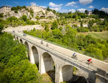 An aerial view of several cyclists crossing a large stone arch bridge above a wooded valley. A historic hilltop village with stone buildings sits on rocky cliffs in the background under a blue sky with scattered clouds.