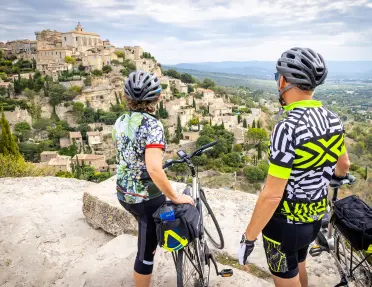 Two cyclists pause at a viewpoint overlooking a dramatic hilltop village built on rocky cliffs. Stone houses, winding streets, and the surrounding countryside stretch into the distance beneath a cloudy sky.