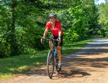 Man smiling while riding a bike on a road covered with leaves, surrounded by tall trees