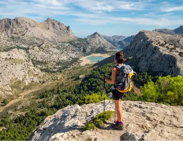 Woman standing on top of a cliff holding two hiking poles, looking down towards tall mountains