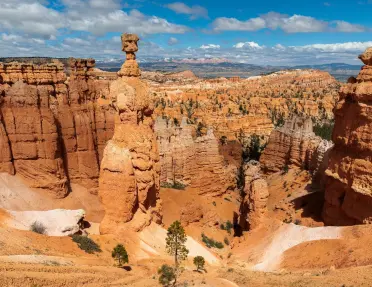 Panoramic view of Bryce Canyon National Park featuring towering orange hoodoos, steep rock formations, and layered desert landscapes beneath a blue sky filled with scattered clouds.