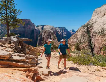 Two hikers walk along a rocky trail in Zion National Park, surrounded by towering sandstone cliffs, canyon walls, and desert vegetation beneath a clear blue sky.